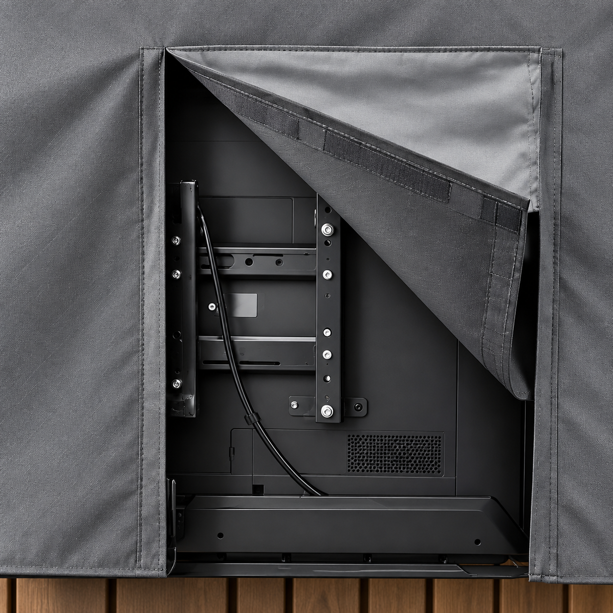 Close-up of a dark gray outdoor TV cover with the front flap pulled open, showing a black television mounted inside. The cover is shown on a wooden deck background.